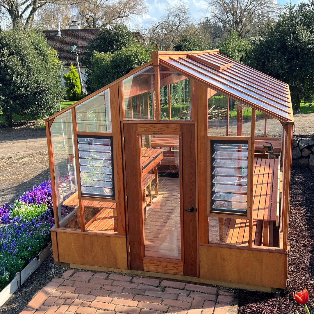 Wooden greenhouse with glass panels on a brick patio, surrounded by greenery.