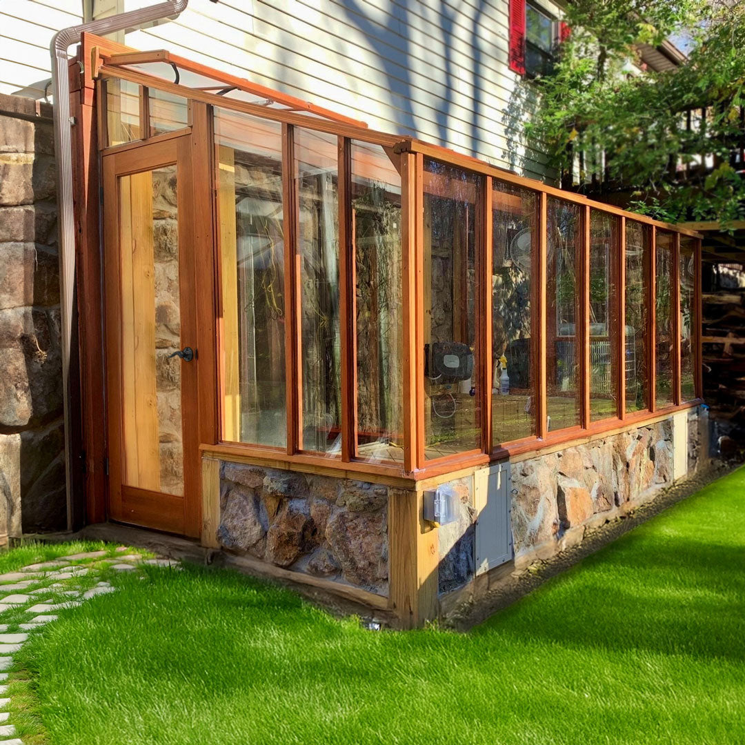 Wooden greenhouse attached to a house with stone foundation, surrounded by green grass.