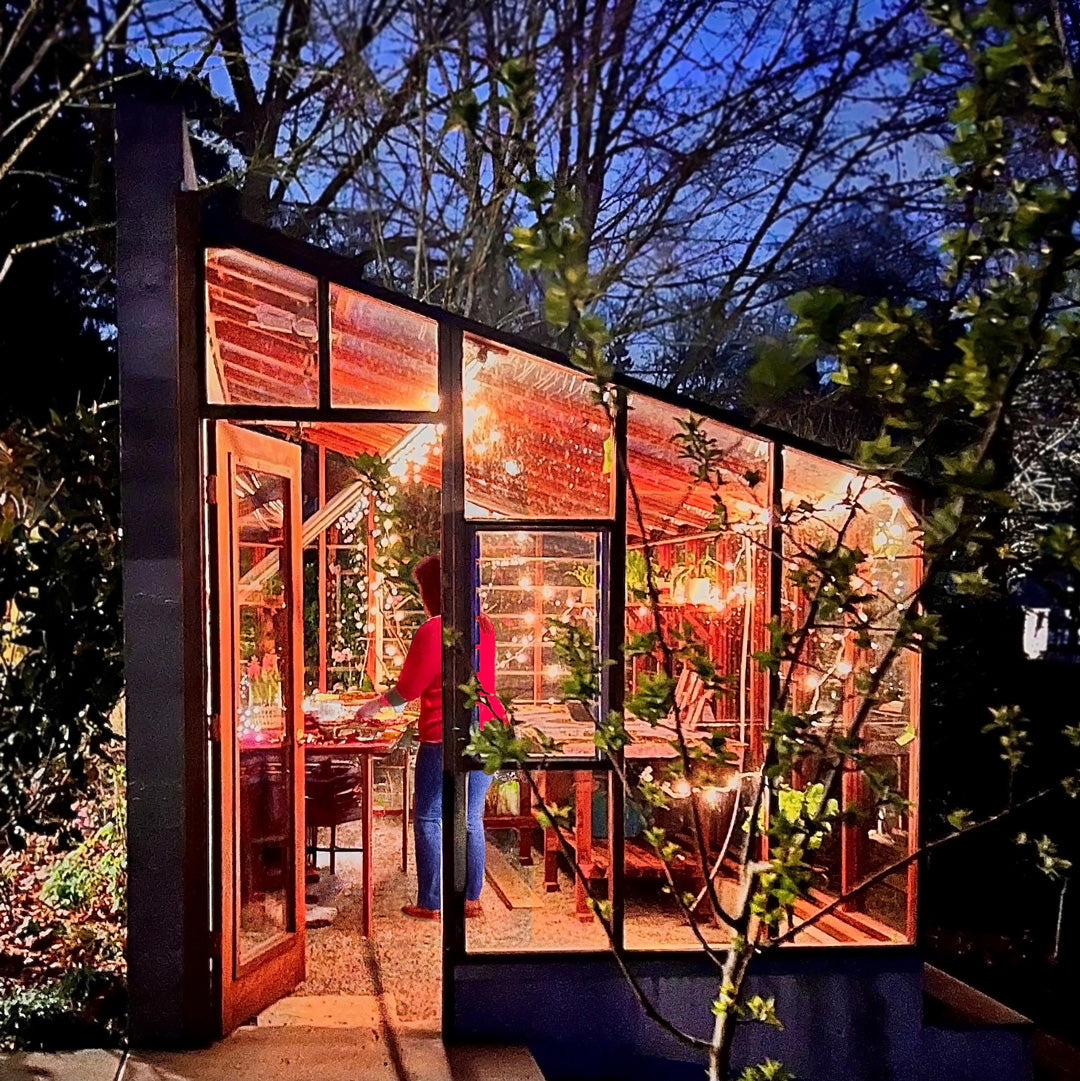 Modern glasshouse with interior lights on, surrounded by trees at night.