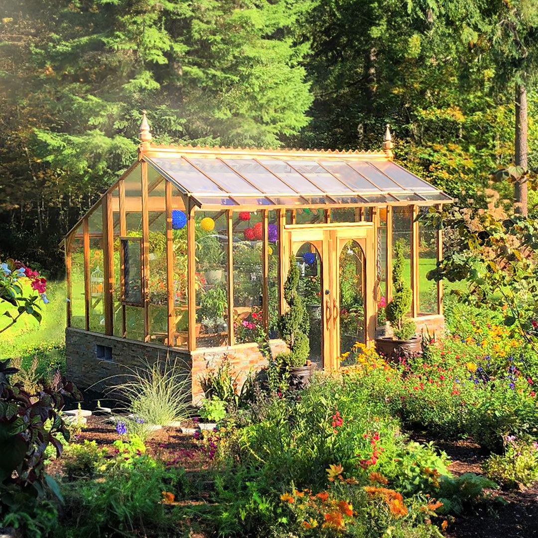 Glass Nantucket greenhouse with brick base in a garden with flowers and trees