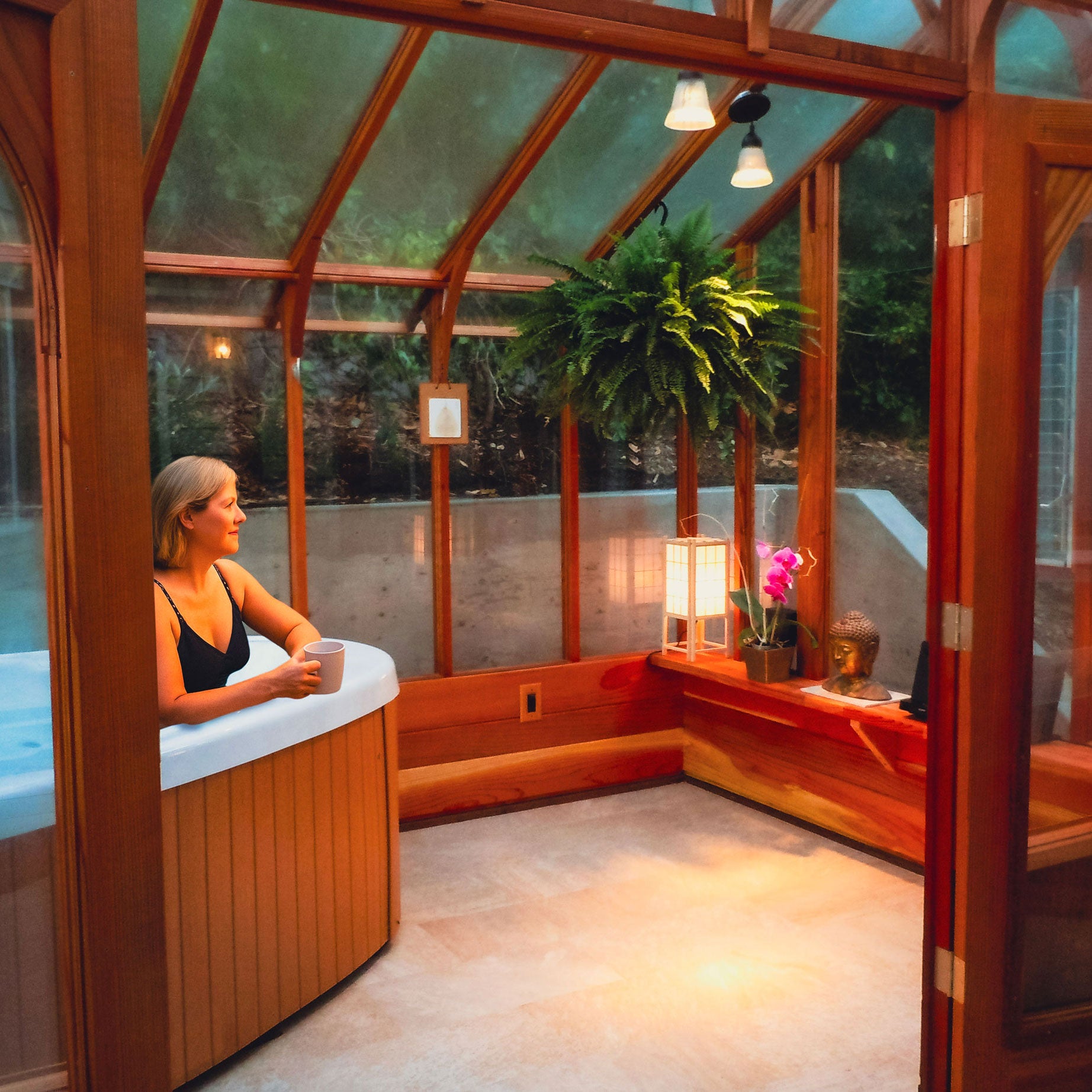 Woman relaxing in a hot tub inside a glass-walled building with plants and decor.