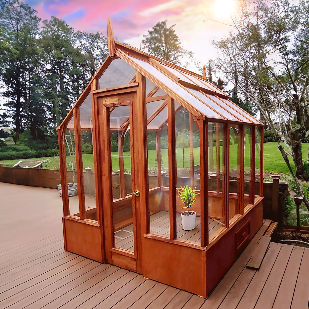 Wooden Trillium greenhouse with glass panels on a wooden deck, surrounded by trees and greenery.
