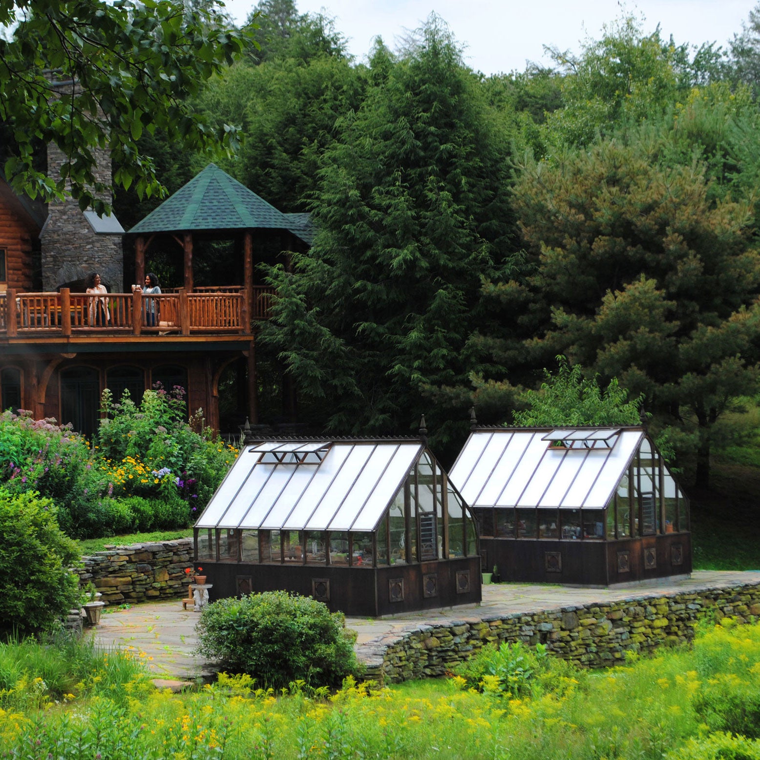 Two greenhouses in a garden setting with a wooden cabin and trees in the background.
