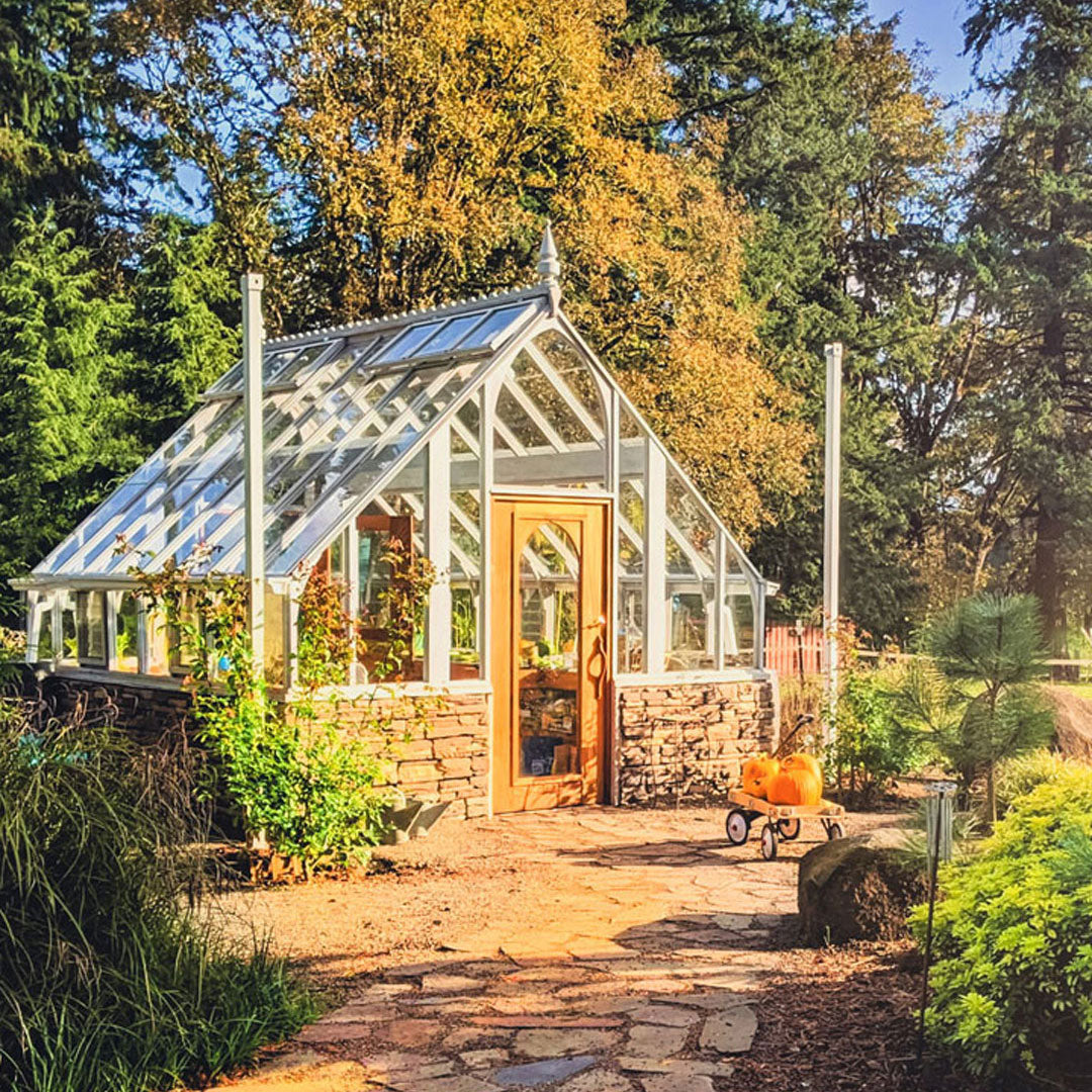 Greenhouse with stone foundation and wooden door surrounded by trees and garden plants.