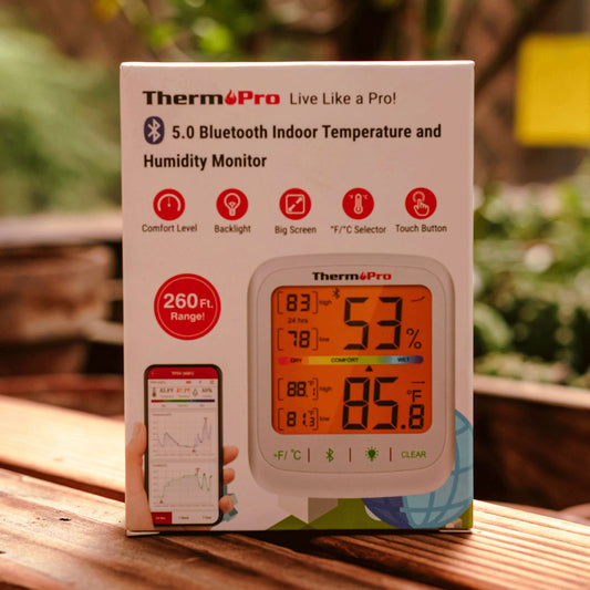 Thermopro temperature and humidity monitor packaging on a wooden surface with blurred greenery in the background