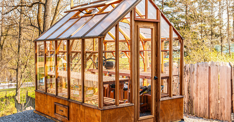 Wood greenhouse with polycarbonate roof and glass walls, nestled in a garden setting
