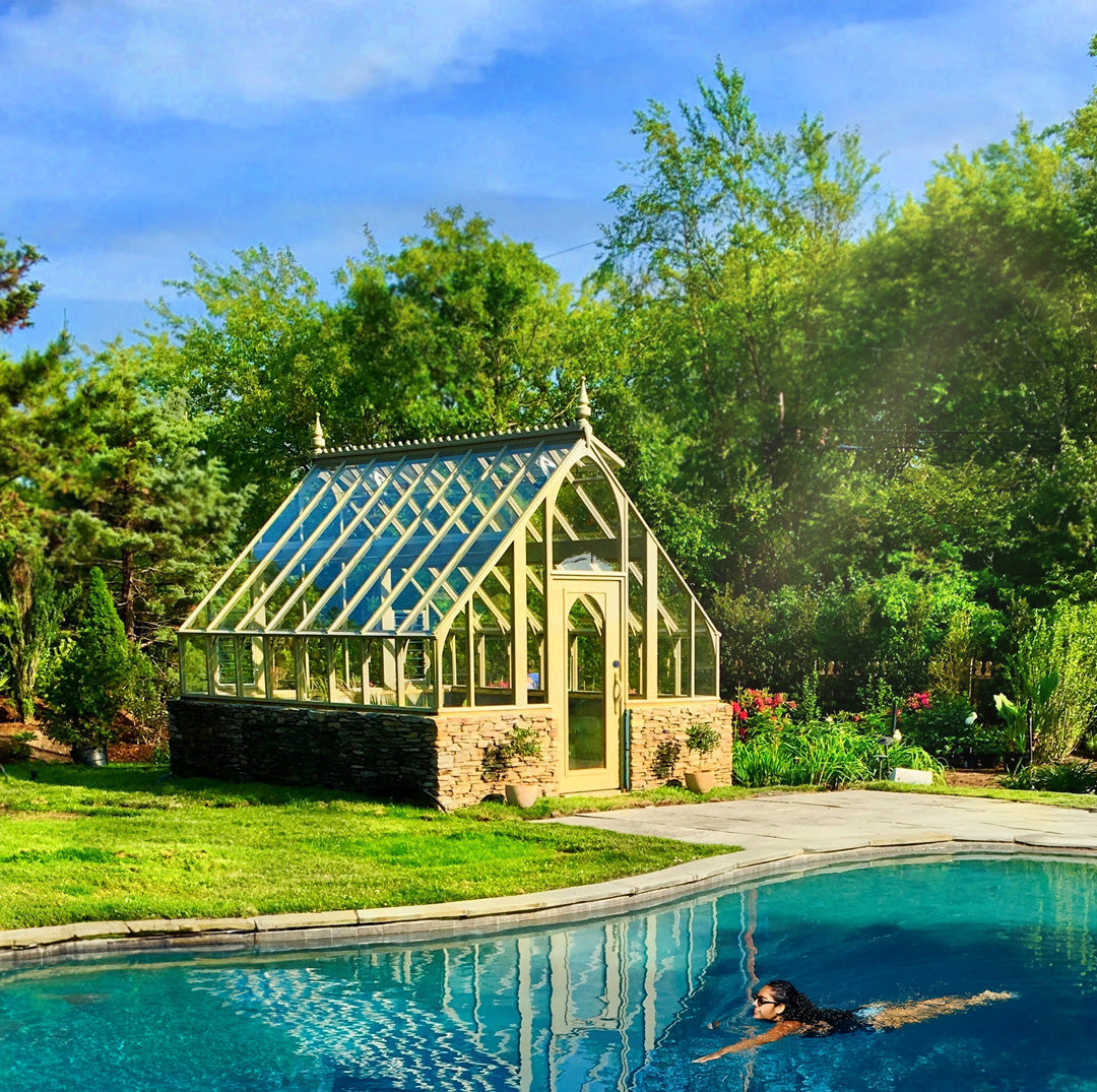 Victorian-style backyard greenhouse with stone base, glass roof, and wood-framed door, surrounded by lush garden landscaping and autumn decor.