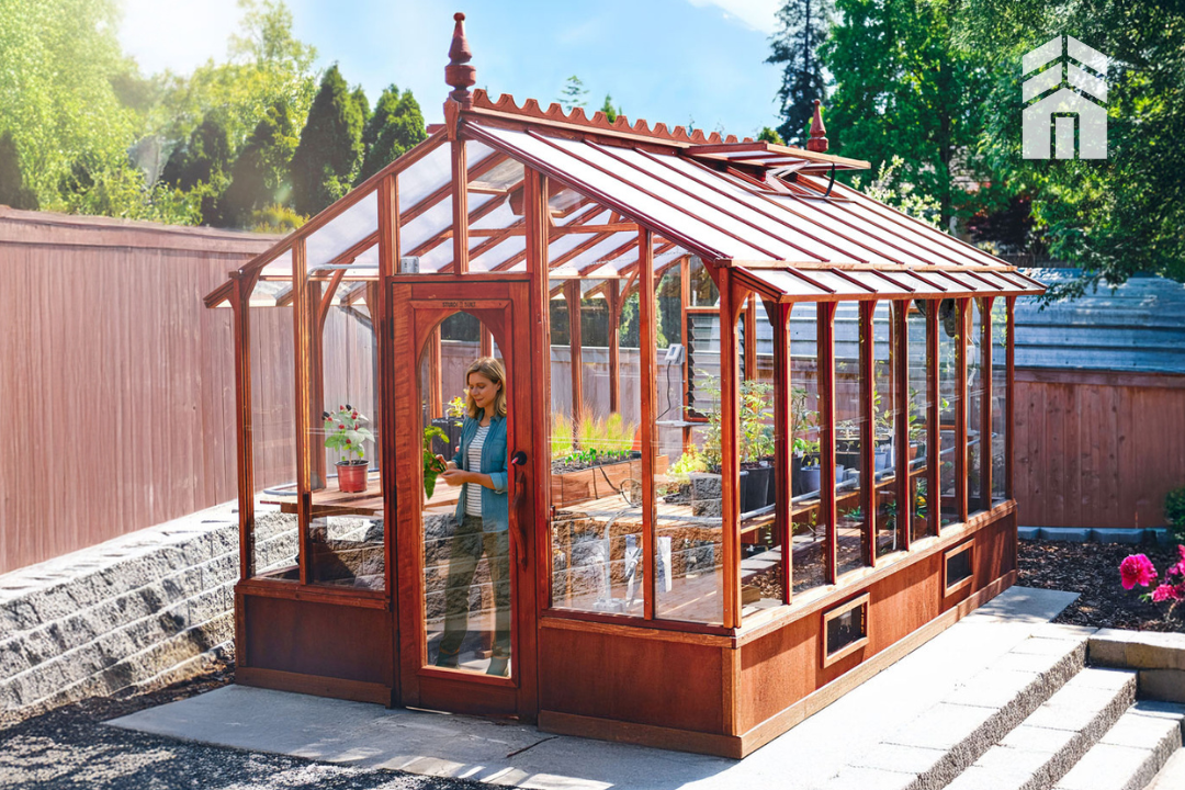 Wooden greenhouse with a person inside, surrounded by plants and garden furniture, on a sunny day.