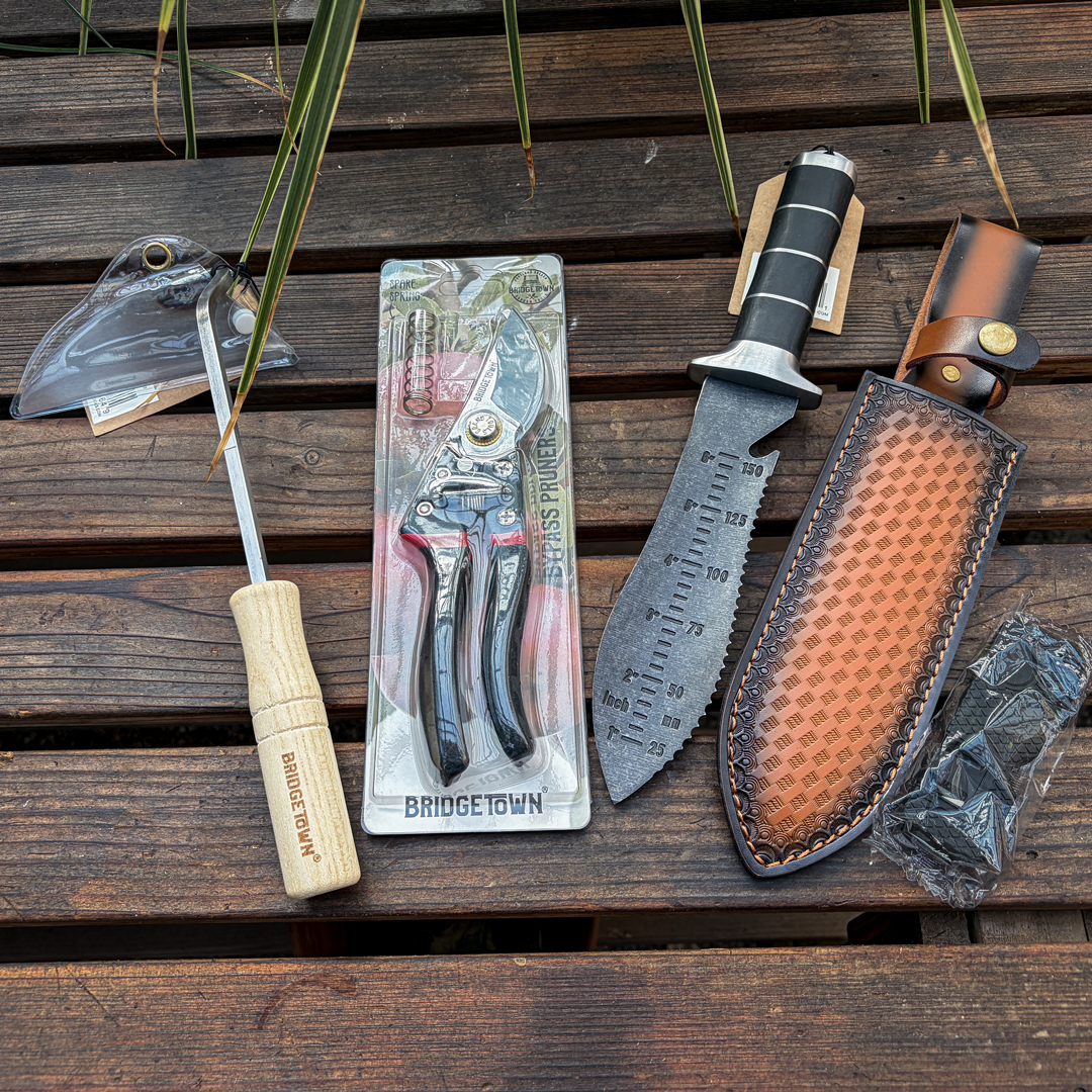A set of outdoor hand tools laid out on a dark wooden surface, including a wooden-handled screwdriver engraved with 'BRIDGE TOWN', a pair of pruning shears in retail packaging, a fixed-blade knife with a black and silver handle, a brown leather sheath, and a small plastic-wrapped accessory.