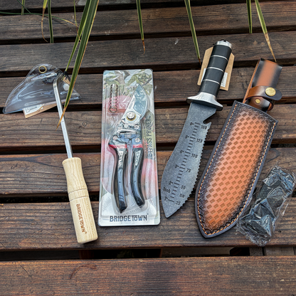 A set of outdoor hand tools laid out on a dark wooden surface, including a wooden-handled screwdriver engraved with 'BRIDGE TOWN', a pair of pruning shears in retail packaging, a fixed-blade knife with a black and silver handle, a brown leather sheath, and a small plastic-wrapped accessory.