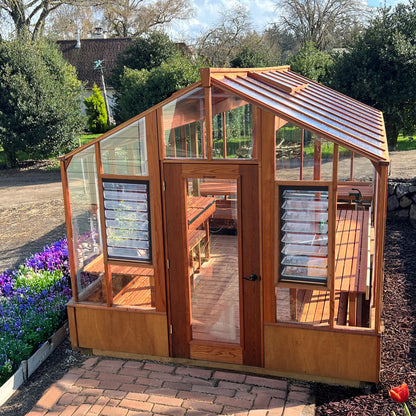 Wooden greenhouse with glass panels on a brick patio, surrounded by greenery.
