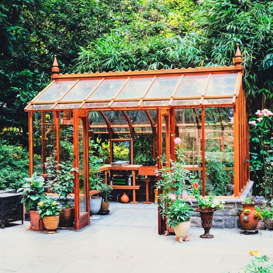 Wooden Nantucket greenhouse with glass panels surrounded by potted plants and trees in backyard garden