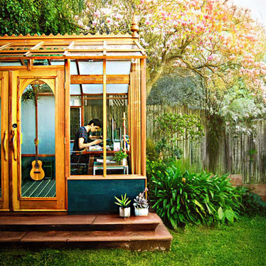 Person inside a wooden shed in a garden with plants and a guitar