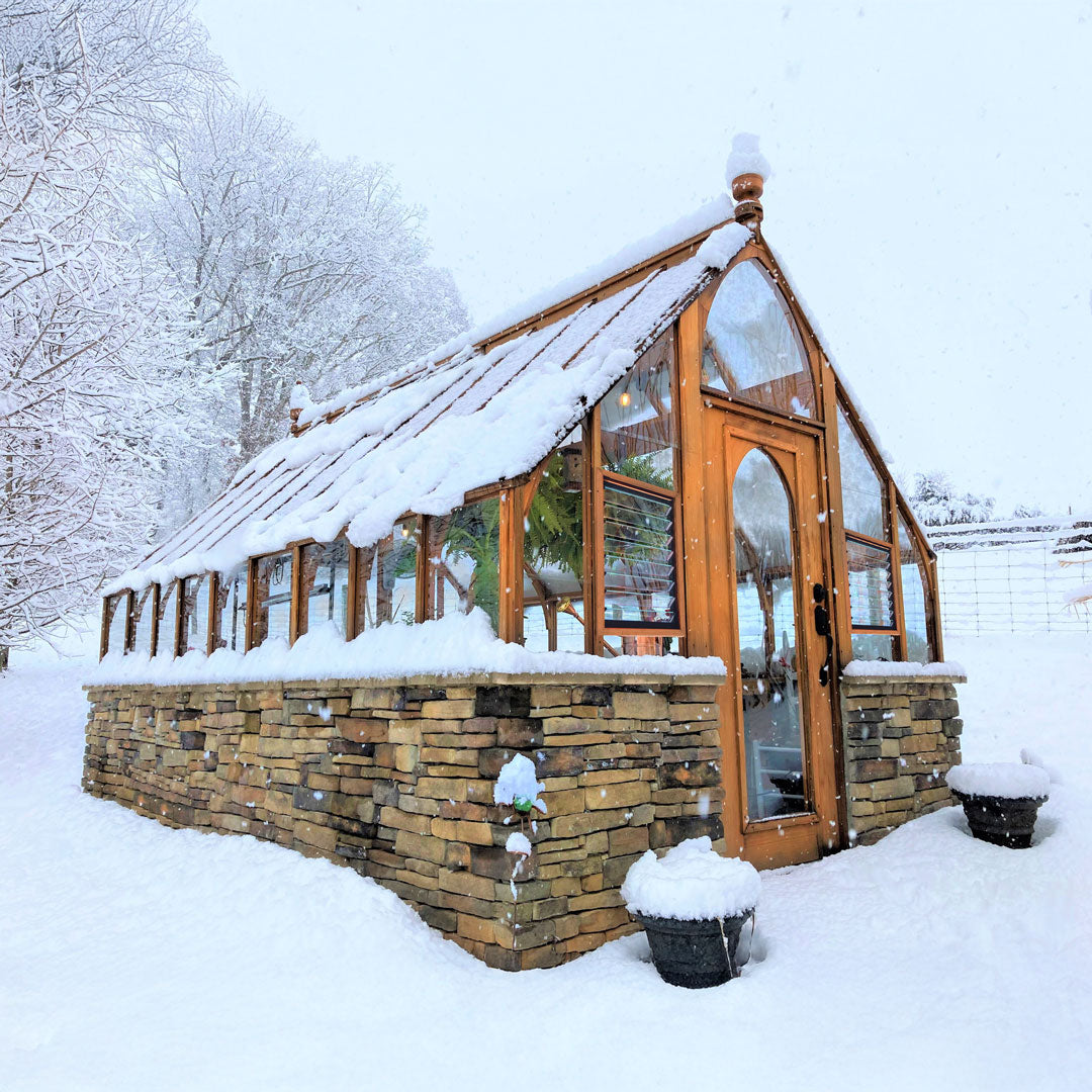 Tudor-style greenhouse in snowy garden, showing steep 12/12 roof and glass panels.